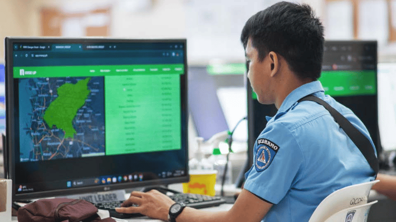 A man in a blue uniform sits at a desk using a computer with a map and data related to mobile-enabled early warning systems displayed onscreen. Office equipment, papers, and another monitor are visible around him in a brightly lit workspace.