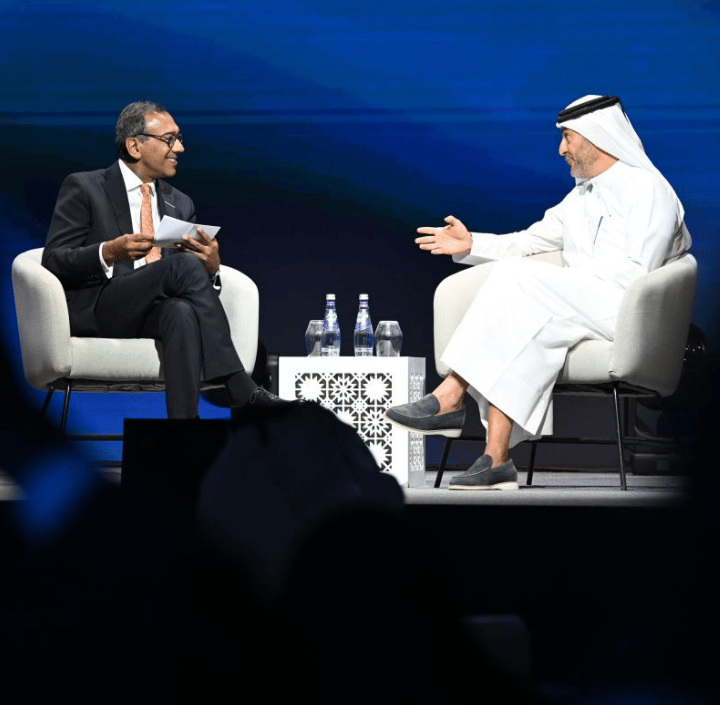 Two men sit in armchairs on a GSMA Mobile for Development stage, facing each other in conversation. On the left, a man in a dark suit holds papers; on the right, a man in a traditional white thobe and ghutra gestures with his hands. They are separated by a small table with water bottles.