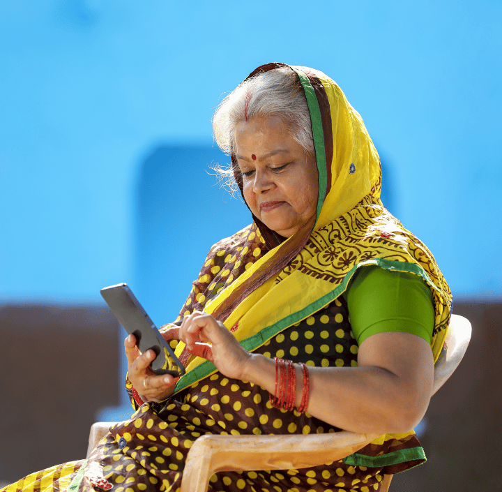 An older South Asian woman with a bindi and grey hair sits against a bright blue wall, wearing a polka-dot sari and red bangles. She looks down at her mobile phone, reflecting the GSMA Mobile for Development focus on digital inclusion.