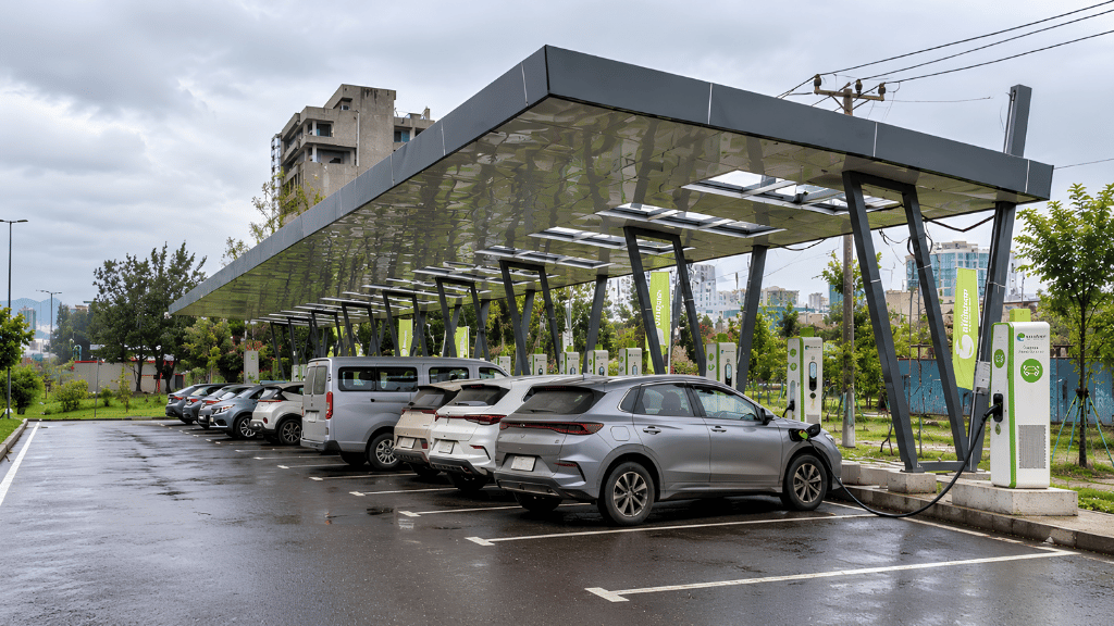 A row of electric vehicles is parked under a modern solar panel canopy at a charging station on a wet, overcast day. Several grey and white SUVs and vans are plugged into tall, green and white charging pillars. In the background, trees and urban buildings appear under a cloudy sky.