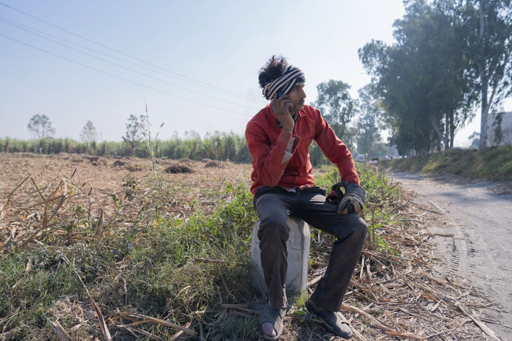 A man in a red shirt and head wrap sits on a concrete post in a field, talking on a mobile phone. He wears dark trousers and work gloves whilst resting beside a dirt path.