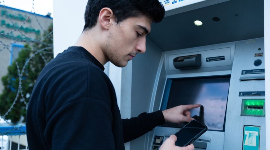 A young man with dark hair wears a black jumper as he interacts with an ATM touchscreen. He holds a mobile phone in his other hand, and in the background, a blue fence topped with barbed wire is visible.