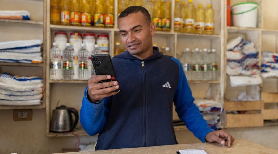 A man with short dark hair wears a blue and navy zip-up hoodie while looking at his mobile phone in a shop. Behind him, wooden shelves are stocked with bottles of cooking oil and various food products.