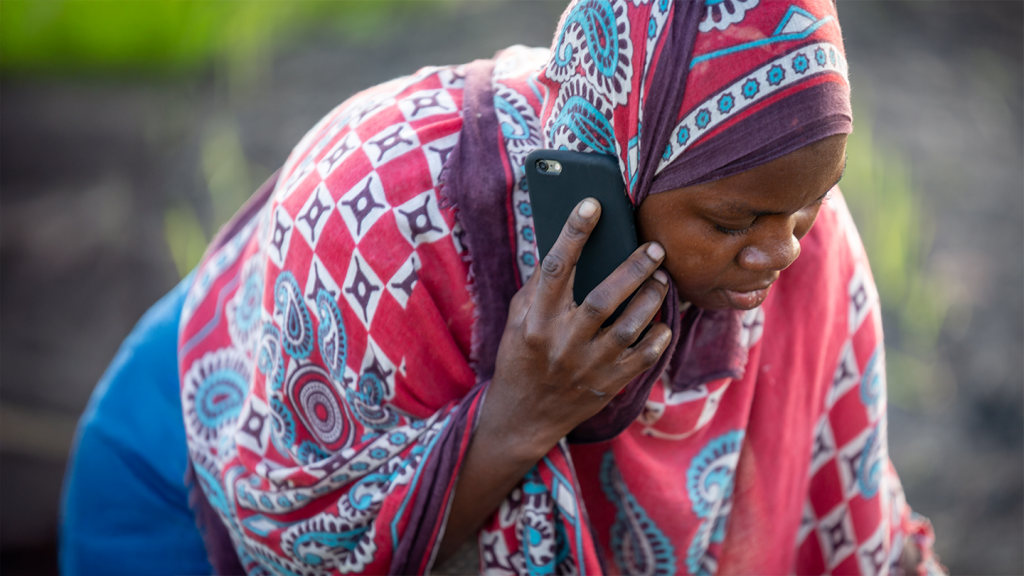 A woman in a colourful red and blue patterned scarf talks on a black mobile phone, reflecting handset affordability, while looking down outdoors with blurred greenery in the background.