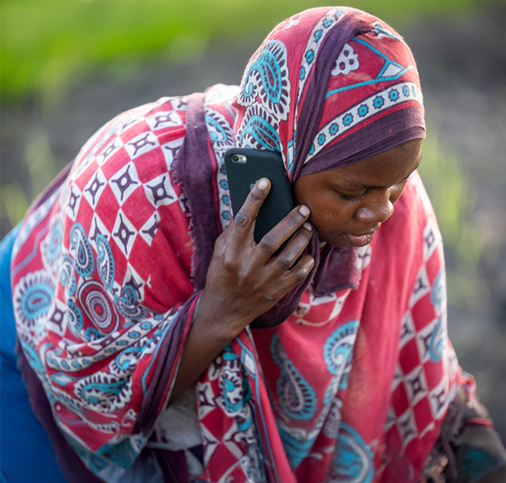 A connected woman wearing a vibrant red and blue patterned headscarf looks down while holding a black mobile phone to her ear. She stands in a field with dark soil, focussing intently on the conversation.