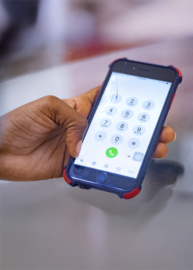 A hand holds a mobile phone with a cracked screen, displaying the phone dialler app. The phone, highlighting handset affordability, is in a blue and red case. The blurred background keeps focus on the hand and device.