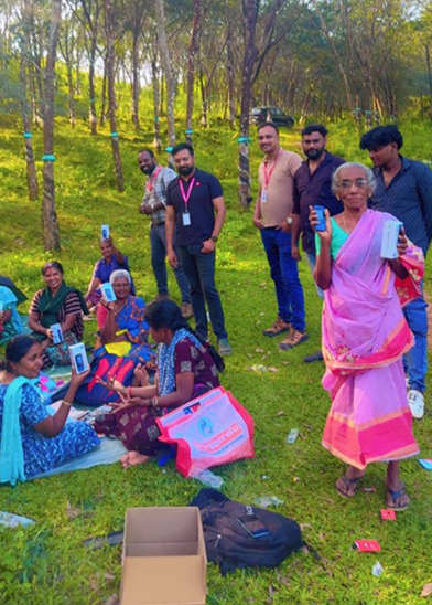 A group of people, some seated on grass and others standing in a lush, sunlit forest. Seated women in colourful sarees proudly display new mobile phones, highlighting handset affordability; nearby, men with lanyards observe the excitement.