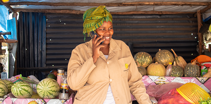 A smiling woman in a beige coat and green headscarf stands at a market stall, talking on her mobile—showcasing how a Connected Society bridges daily life and technology amid pumpkins, watermelons, and a simple corrugated backdrop.