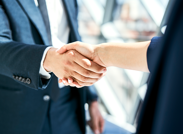 Two people in business attire shake hands indoors, signifying agreement or partnership on a Mobile Policy Handbook. Only their hands and part of their torsos are visible. The background is bright, with large glass windows and blurred architectural details.