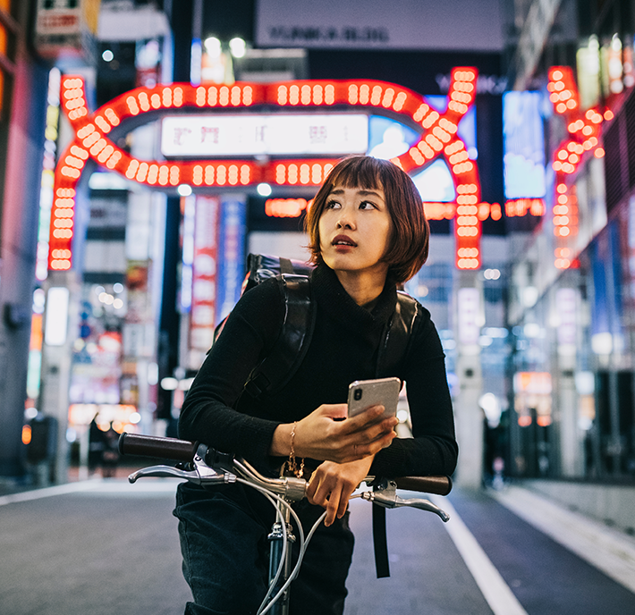 A person with a bob haircut, holding a smartphone, looks away thoughtfully while leaning on a bicycle under neon signs at night in an urban setting.