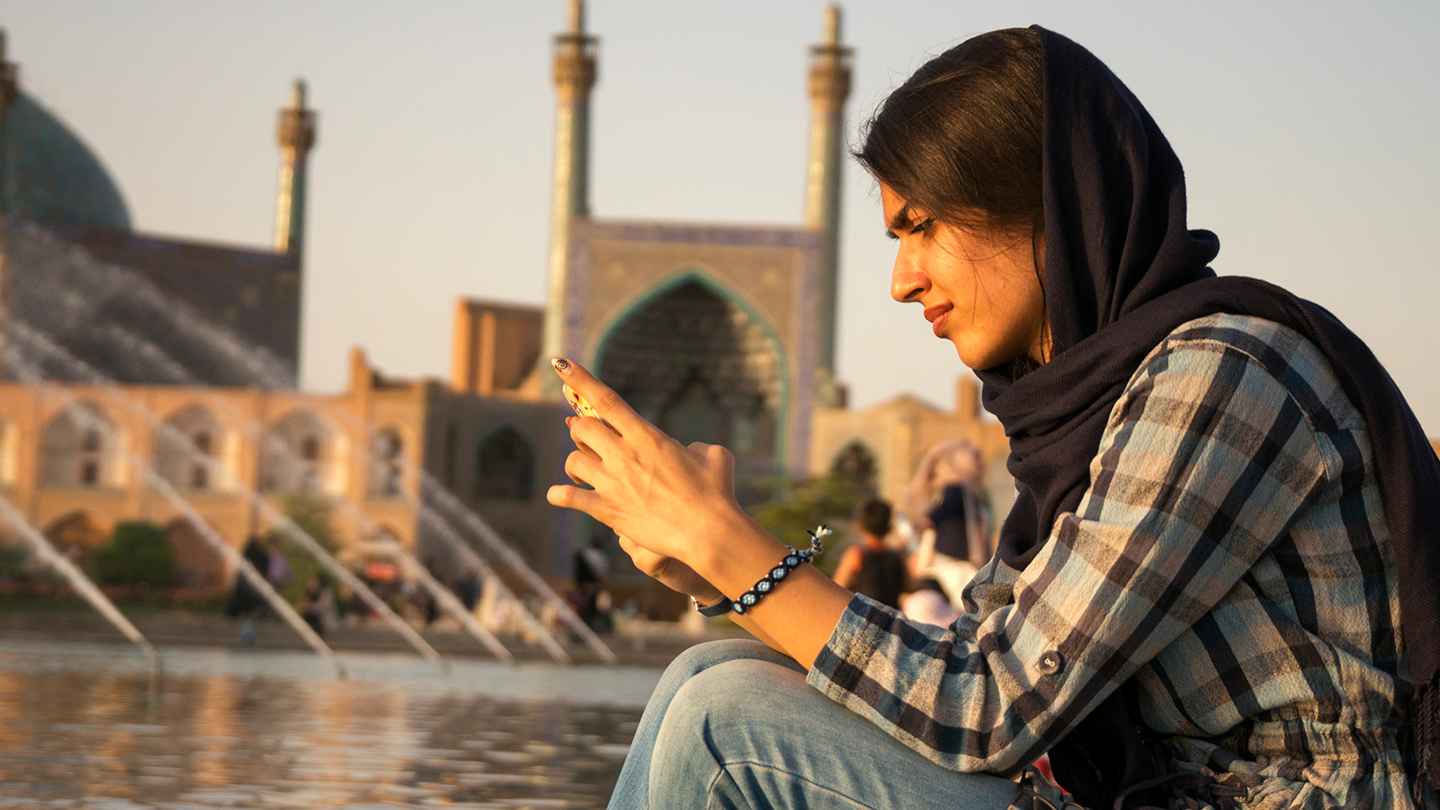 A person wearing a headscarf sits by a reflective pool, looking at her smartphone, with a mosque in the background at sunset.