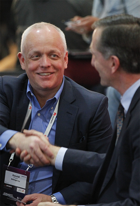 Two people in business attire, one bald and one with dark hair, shaking hands and smiling during a conversation at a conference.