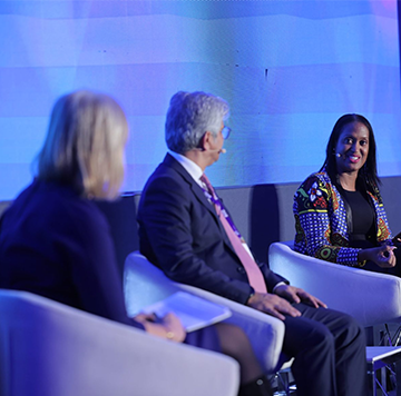 Three professionals engaged in a discussion on stage at a conference, with a person in a colorful blazer speaking to two colleagues.