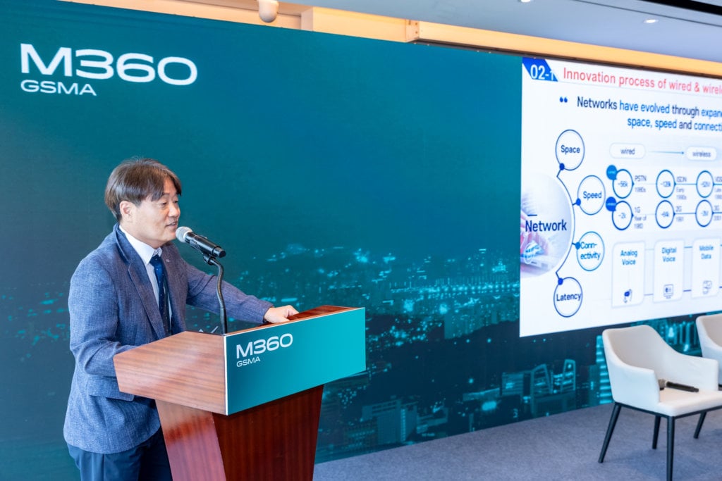 A man in a suit stands at a podium with a microphone, speaking at an event. The background shows a large screen displaying a presentation slide titled "Innovation process of wired & wireless." The wall and podium feature the "M360 GSMA" logo.