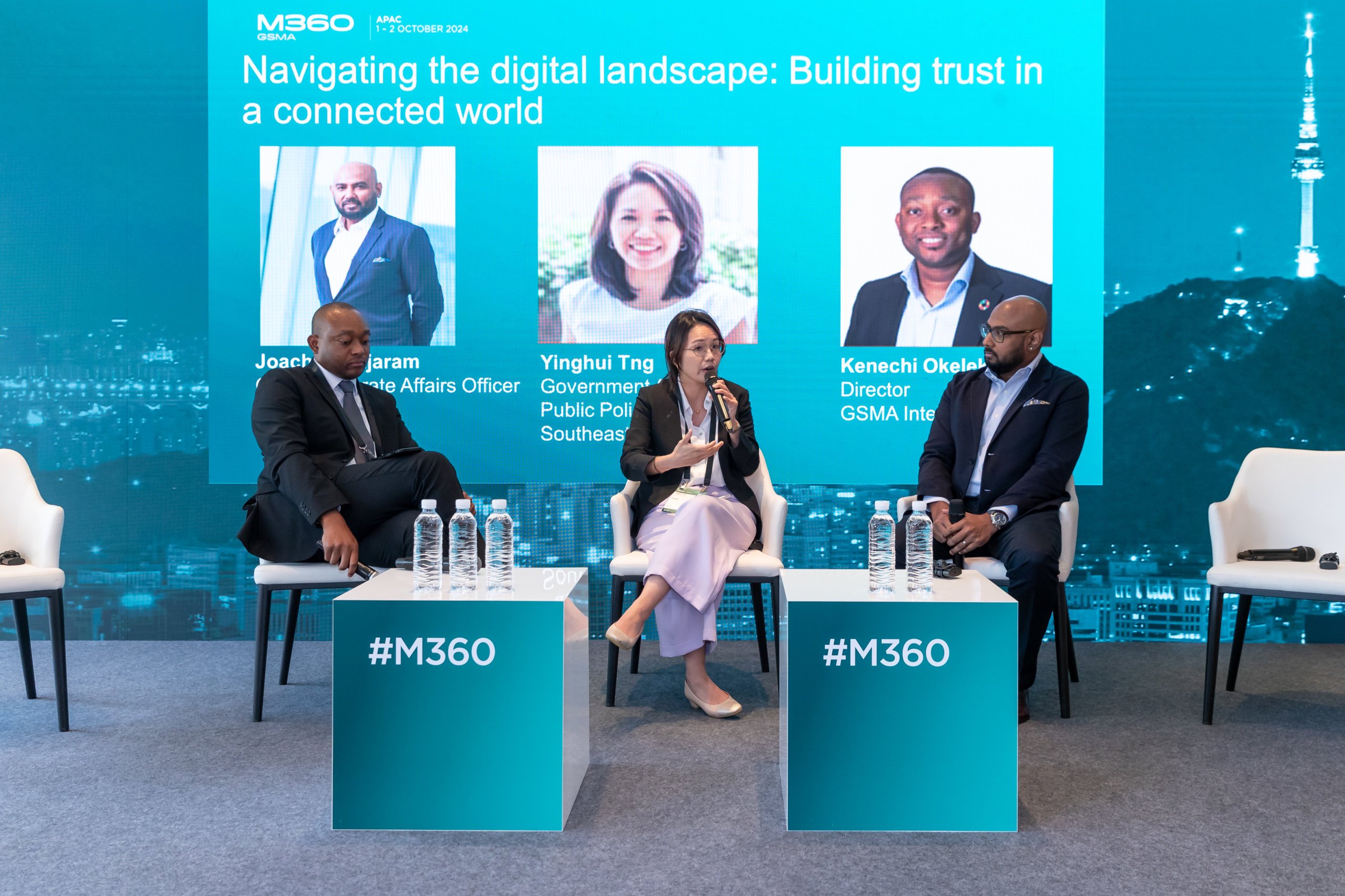 Three individuals are seated on a stage during a panel discussion titled "Navigating the digital landscape: Building trust in a connected world." A backdrop displays panelists’ names and affiliations. Bottled water is on a small table in front of them.