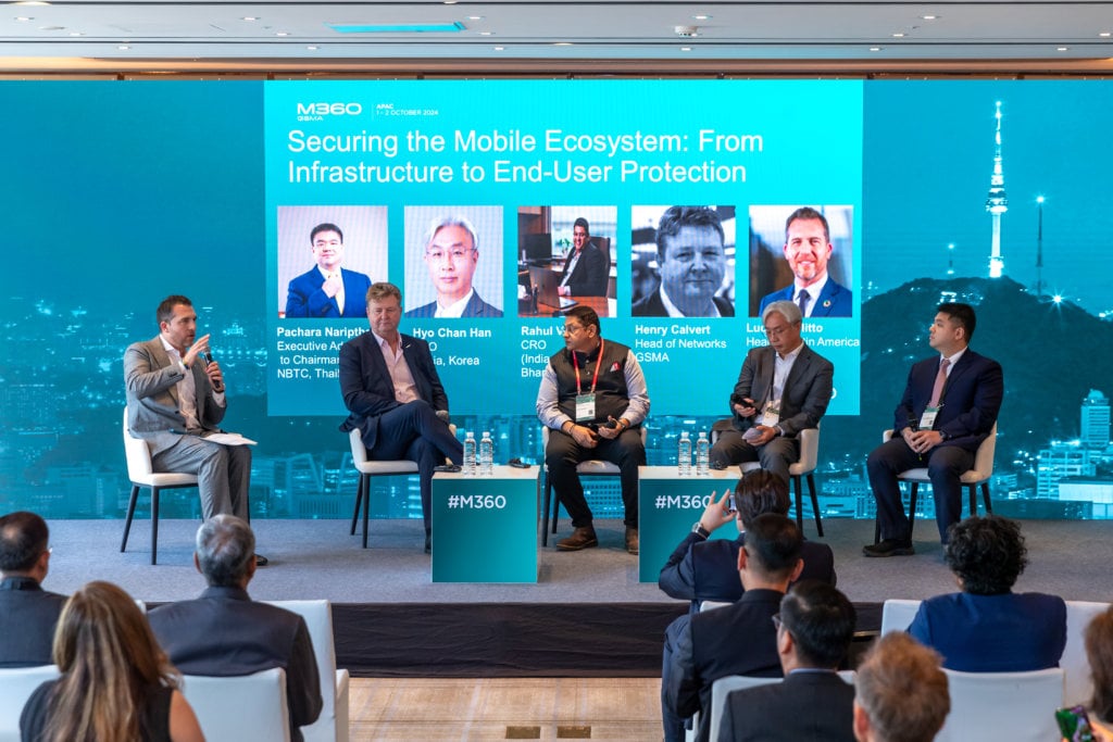 A panel of five people sit on stage in front of a screen displaying the text "Securing the Mobile Ecosystem: From Infrastructure to End-User Protection." An audience is seated in front of them.