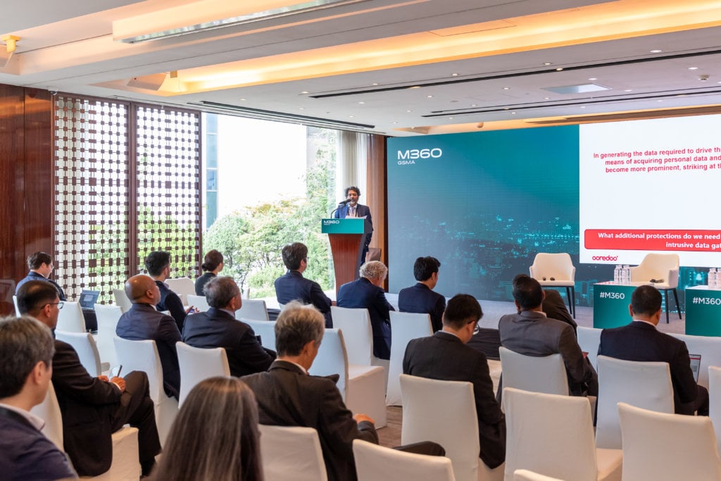A speaker is presenting at a conference in a modern, well-lit room. Attendees are seated facing the speaker and the presentation screen. The screen displays a presentation slide with text. A sign reads "M360" near the platform where the speaker stands.