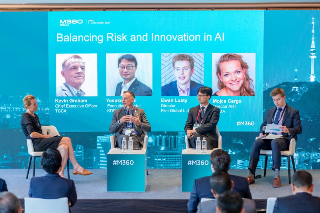A group of five people are seated on a stage with a large screen behind them displaying, "Balancing Risk and Innovation in AI." Four of them are in chairs on stage, and one is standing, moderating the discussion. An audience is visible in the foreground.