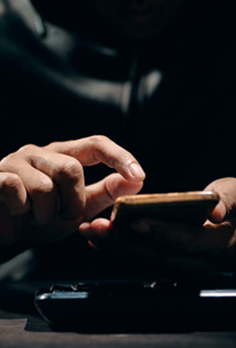 In a dimly lit setting, an individual uses a smartphone, one hand holding it while the other taps the screen. The shadows obscure their face, spotlighting their hands and phone. This intimate moment echoes the connectivity goals championed by GSMA Public Policy initiatives.