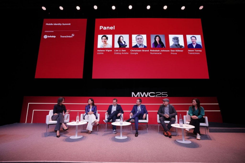 A panel of six people sits on a stage with red and white decor at the Mobile Identity Summit. A large screen above displays their images and names. The stage flooring has "MWC23" written on it. The panelists are engaged in discussion, each seated on a white chair with small tables in front.