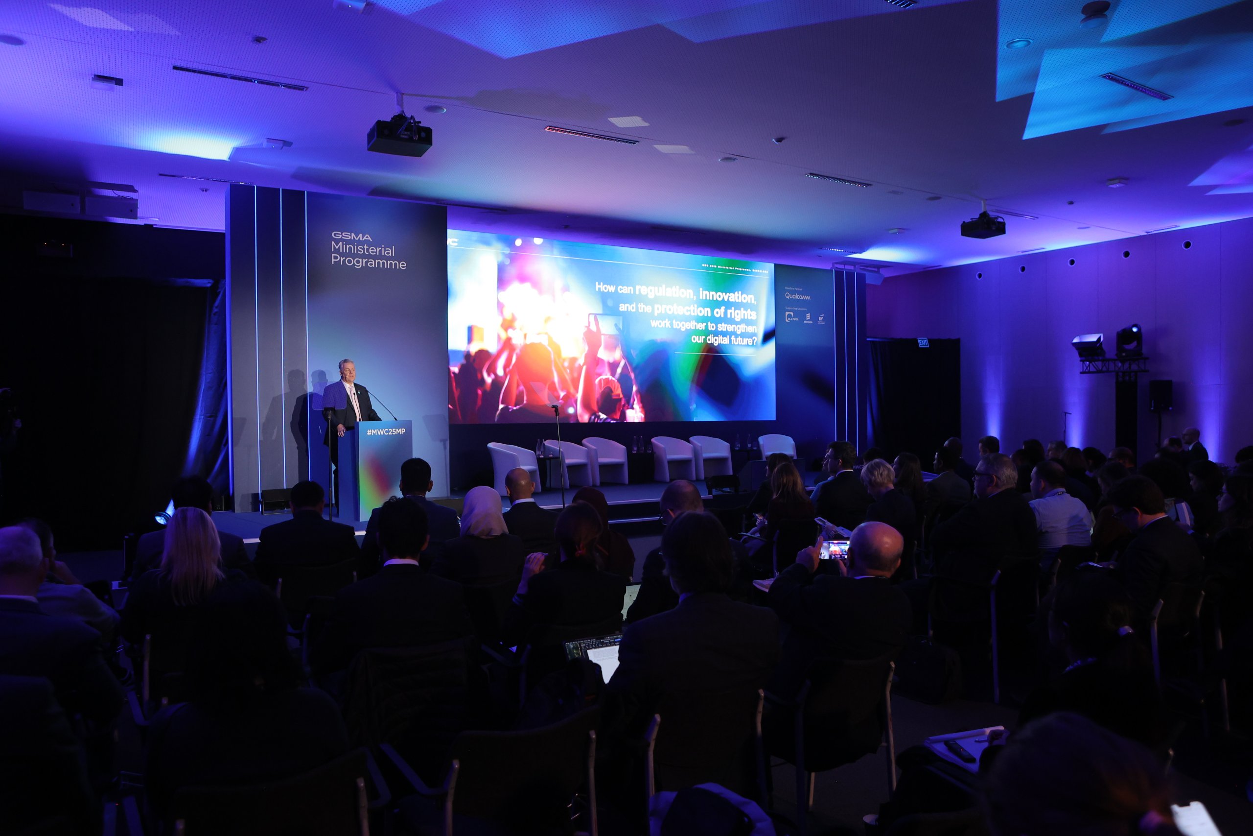 A speaker addresses an audience at a conference. The stage has blue lighting and a large screen displaying a presentation slide about regulation and rights. Four white chairs are arranged on stage for a panel discussion. The audience is seated, some using laptops and taking notes.