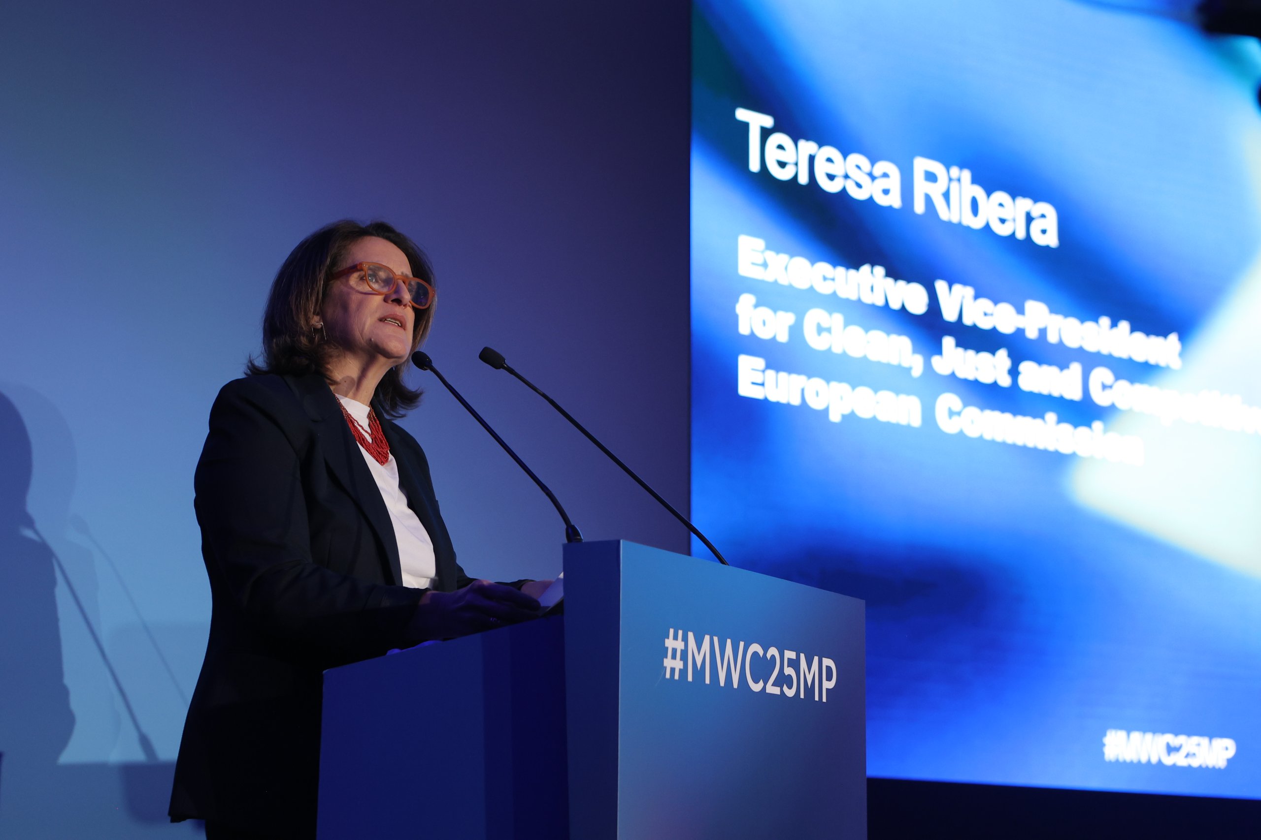 A person in a dark suit and glasses speaks at a podium with microphones. The podium is labeled "#MWC25MP." A screen behind displays text: "Teresa Ribera, Executive Vice-President for Clean, Just and Competitive, European Commission." The setting appears to be a conference or event.