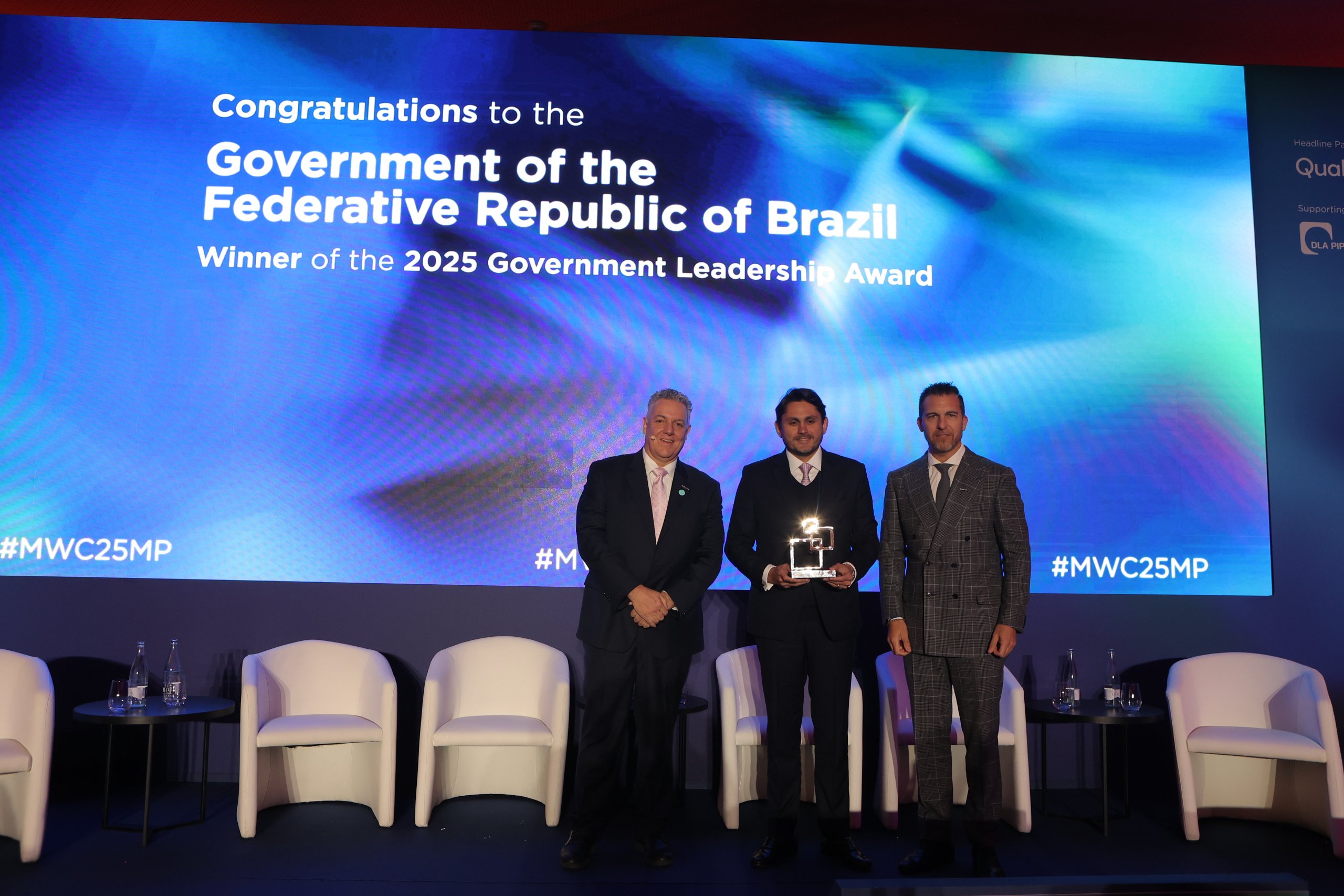 Three individuals stand on stage holding an award, with a backdrop screen displaying "Congratulations to the Government of the Federative Republic of Brazil. Winner of the 2025 Brazil Government Leadership Award." Two white chairs are visible in the background.