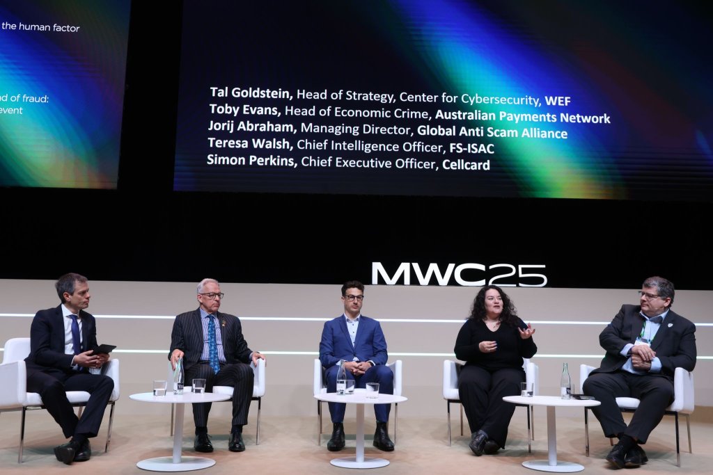 A panel of five people seated on a stage with a dark backdrop displaying their names and titles, discussing cybersecurity. The stage features "MWC25" in white text. The panelists, in business attire, are engaged in conversation, with some gesturing as they speak.