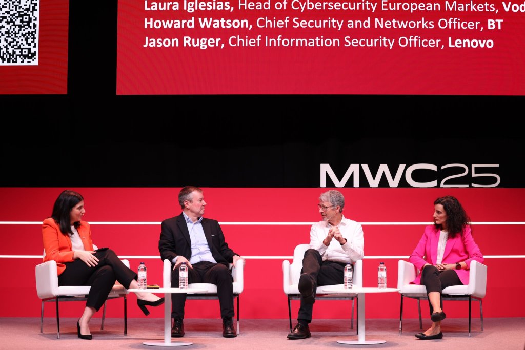 Four panelists are seated on stage at the MWC25 event, beneath a screen displaying panelist names and titles. They are engaged in discussion, with water bottles on the small tables between them. The backdrop is red and black, and an audience member is partially visible in the foreground.