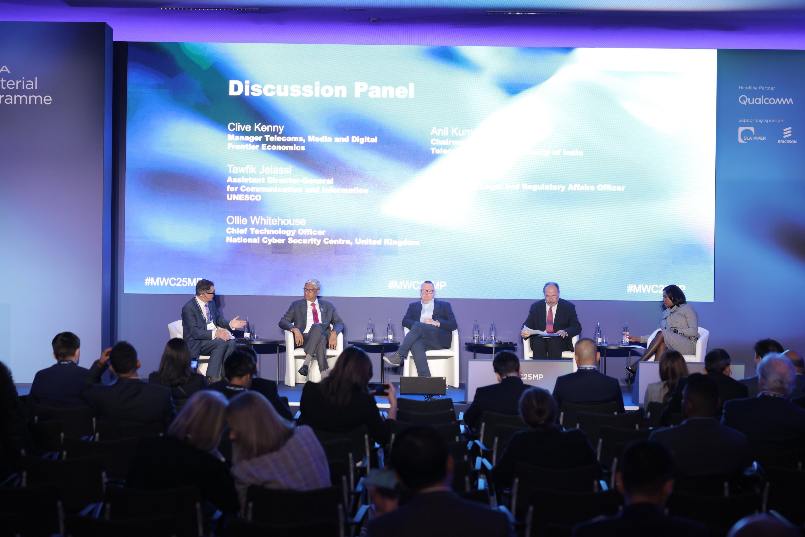 A discussion panel at a conference features five speakers seated on a stage. They are engaged in conversation. Behind them is a large screen displaying their names and roles. The audience is seated in the foreground. The lighting is dim, with a blue and white color scheme.