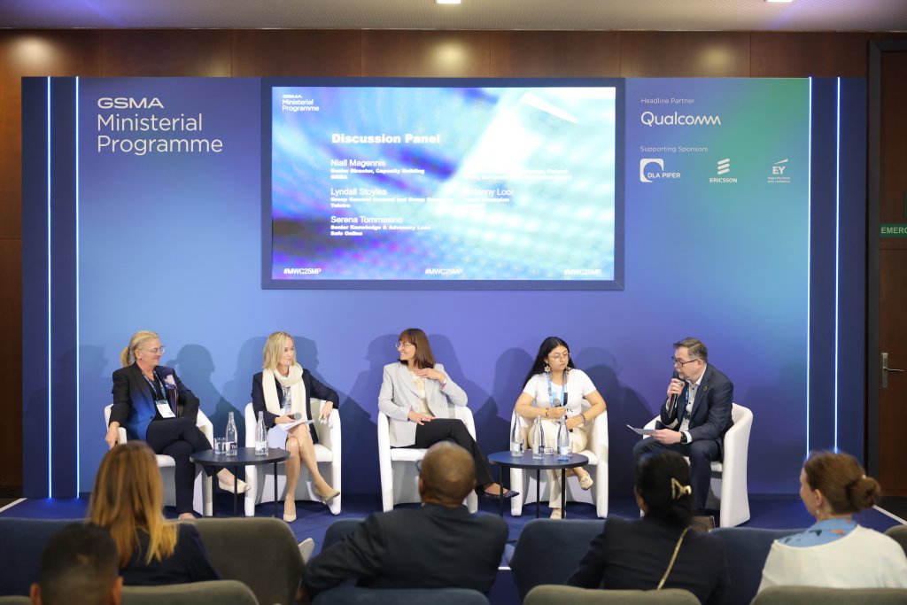 A panel discussion at a conference with five seated participants on a stage. Three women and two men are talking, with microphones and notes in hand. A large digital screen behind them displays the event title and sponsors. Audience members are visible in the foreground.