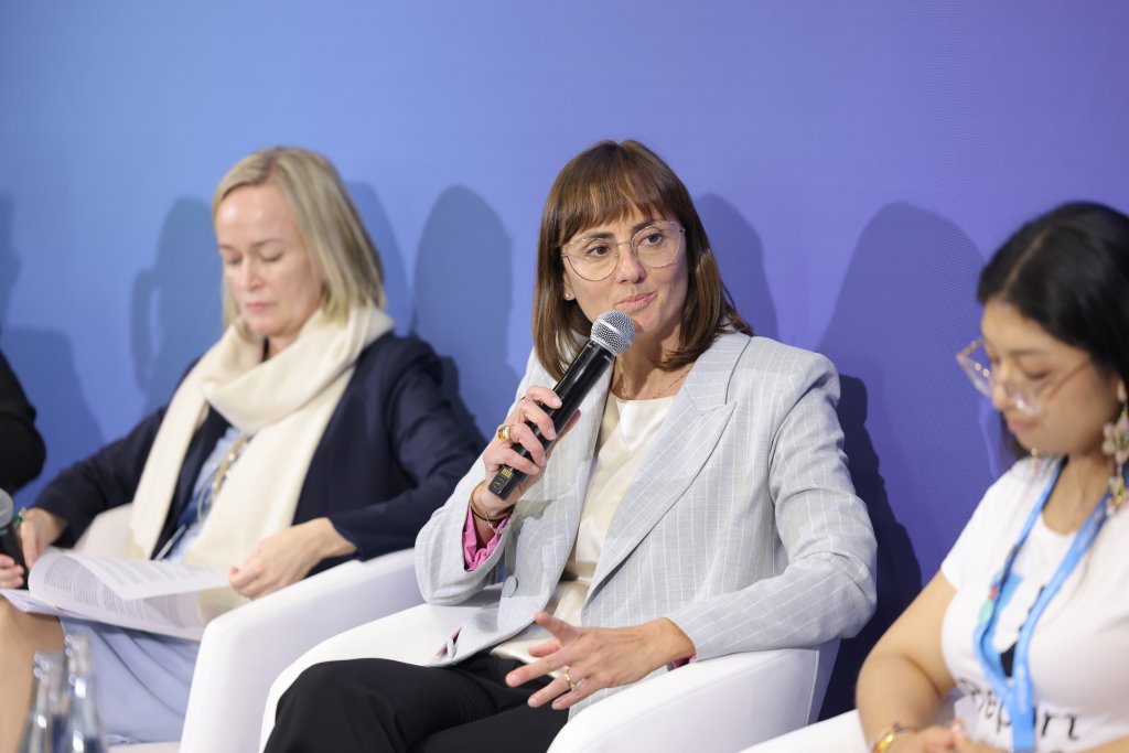 Three women are seated on a panel against a blue background. The center woman, wearing glasses and a light gray blazer, speaks into a microphone. The woman on the left is reading documents, wearing a dark blazer and light scarf. The woman on the right wears a white T-shirt and glasses.