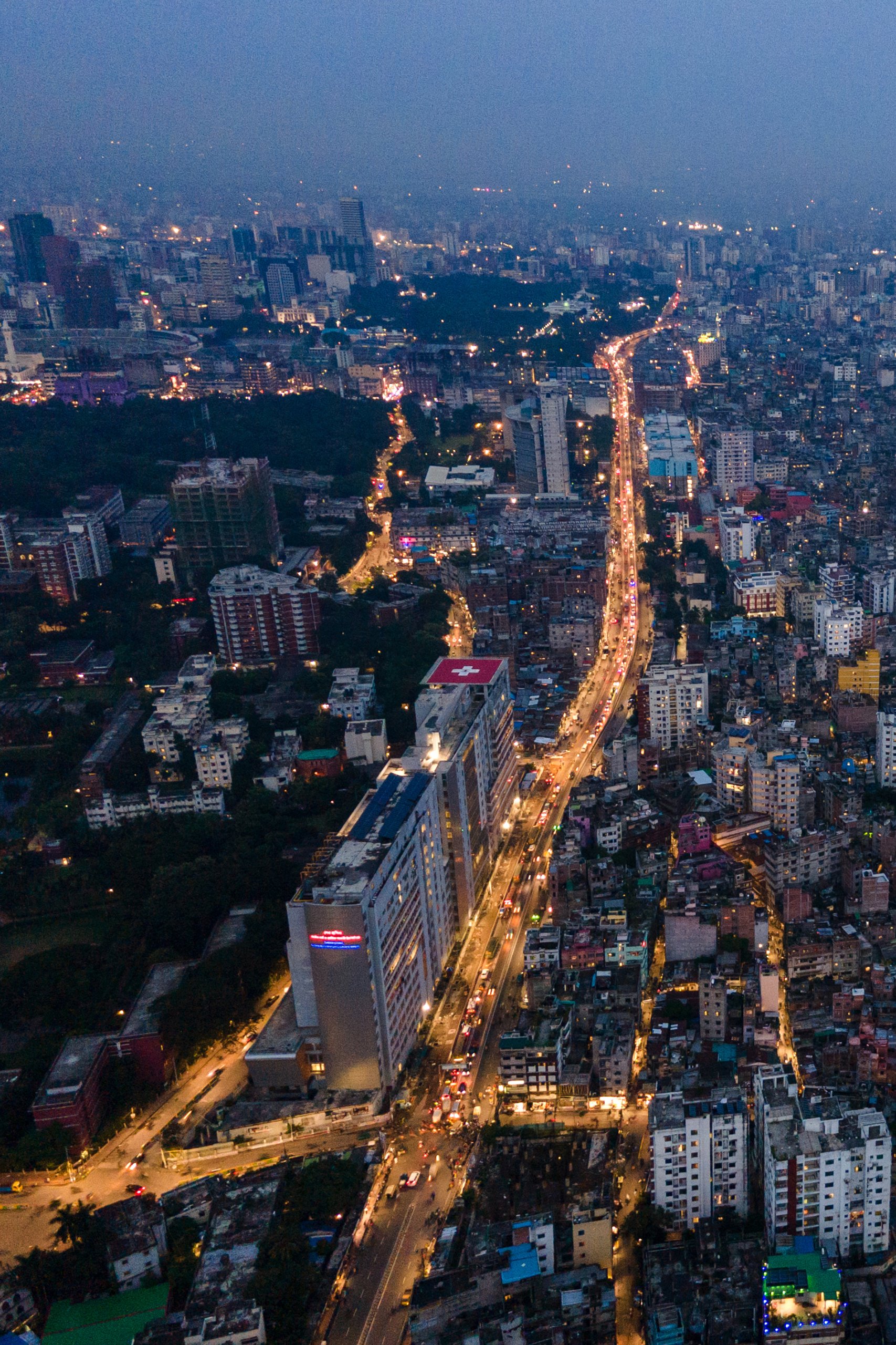 Aerial view of a city at dusk with a long, winding road lit by streetlights cutting through the densely packed buildings. Tall modern structures rise prominently, echoing GSMA Public Policy’s vision for urban connectivity. The background fades into a misty horizon, showcasing the city's vastness.
