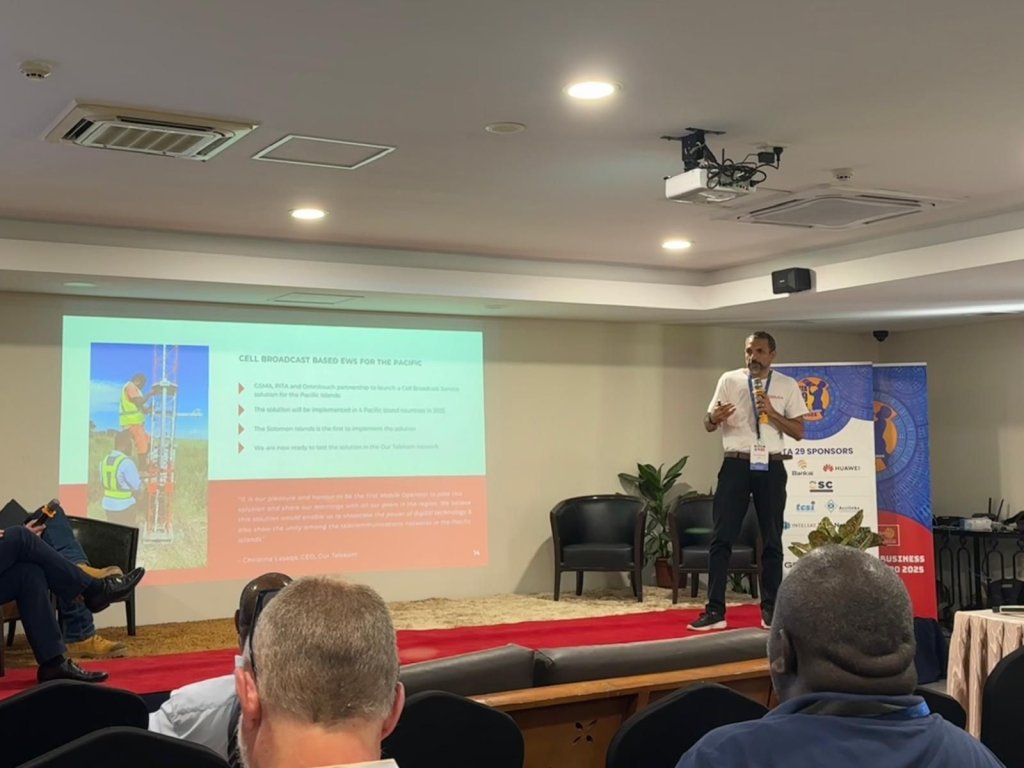 A man gives a presentation onstage in a conference room, discussing digital transformation in the Pacific. A slide with two workers at a cell tower is projected, while attendees and conference banners are visible in the background.