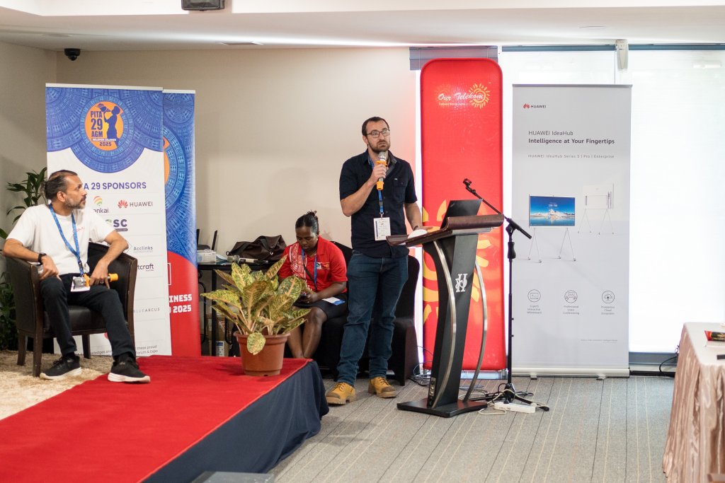 A man stands at a podium speaking about digital transformation in Pacific regions at a conference. Two people sit nearby; one on stage, one beside the podium. Sponsor banners, a red carpet, a potted plant, and large windows brighten the setting.