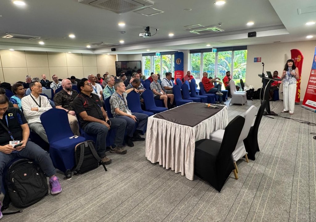 A woman stands and presents at a podium in a conference room, discussing digital transformation in Pacific. Attendees, a mix of men and women, sit attentively in blue chairs. Banners and windows with greenery set a formal scene.