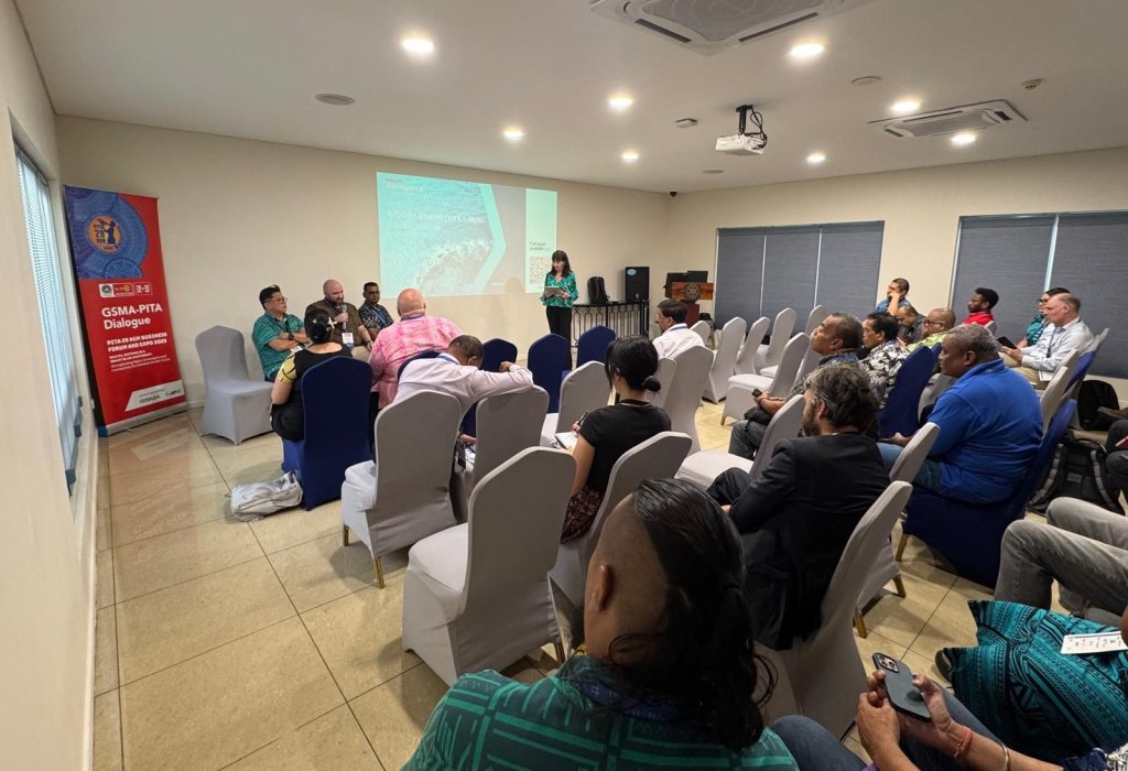 A group of people sit on chairs facing a panel discussion in a small conference room. One person stands and speaks at the front near a screen presenting on digital transformation in Pacific. Attendees listen attentively; banners and closed blinds are visible behind them.