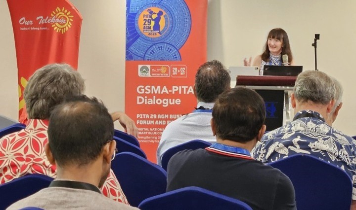 A woman stands at a podium speaking to an audience about digital transformation in Pacific communities. Behind her, red banners read "GSMA-PITA Dialogue" and "PITA 29 AGM 2025," as several attendees in patterned shirts listen attentively from blue chairs.