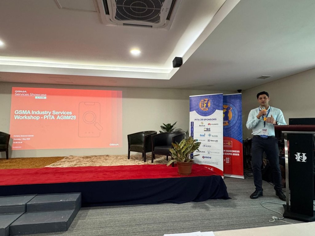 A man in business attire stands at a podium speaking into a microphone in a conference room. Behind him, a projector screen displays a red slide titled "GSMA Industry Services Workshop - PITA AGM#29" on digital transformation in Pacific.