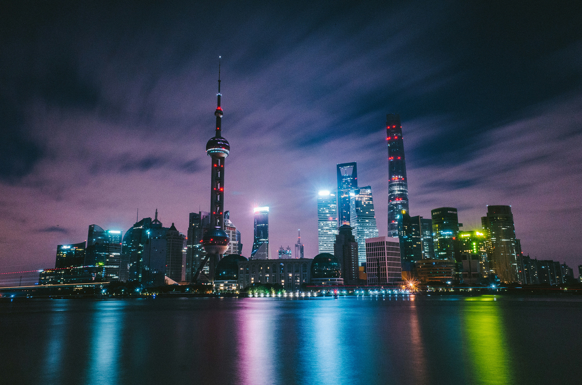 A nighttime cityscape of Shanghai with illuminated skyscrapers reflecting on the Huangpu River under a cloudy purple sky. The Oriental Pearl Tower stands out among other tall buildings, displaying vibrant lights and creating a striking urban skyline.