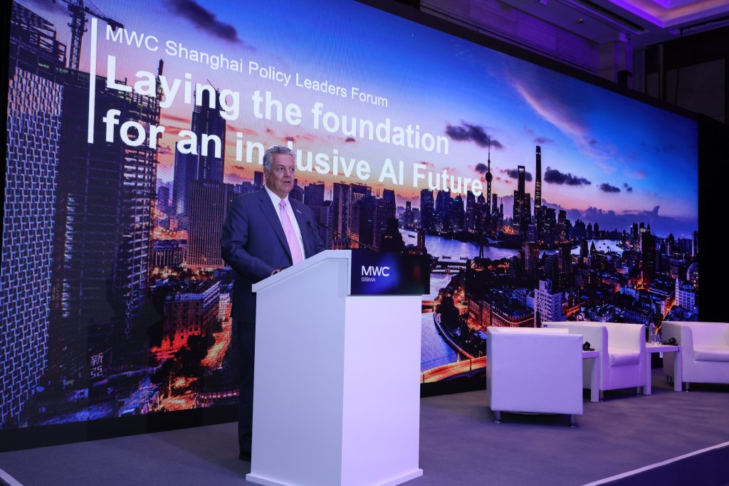 A man in a suit stands at a podium labeled "MWC" on a stage, speaking at the MWC Shanghai Policy Leaders Forum. Behind him is a large screen displaying a city skyline at sunset and text reading "Laying the foundation for an inclusive AI Future.