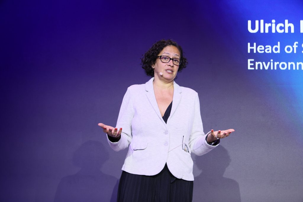 A woman with curly hair and glasses, wearing a white blazer over a dark top, stands onstage with a headset microphone. She gestures with both hands, palms up, in front of a purple-lit background featuring partial text on the right.