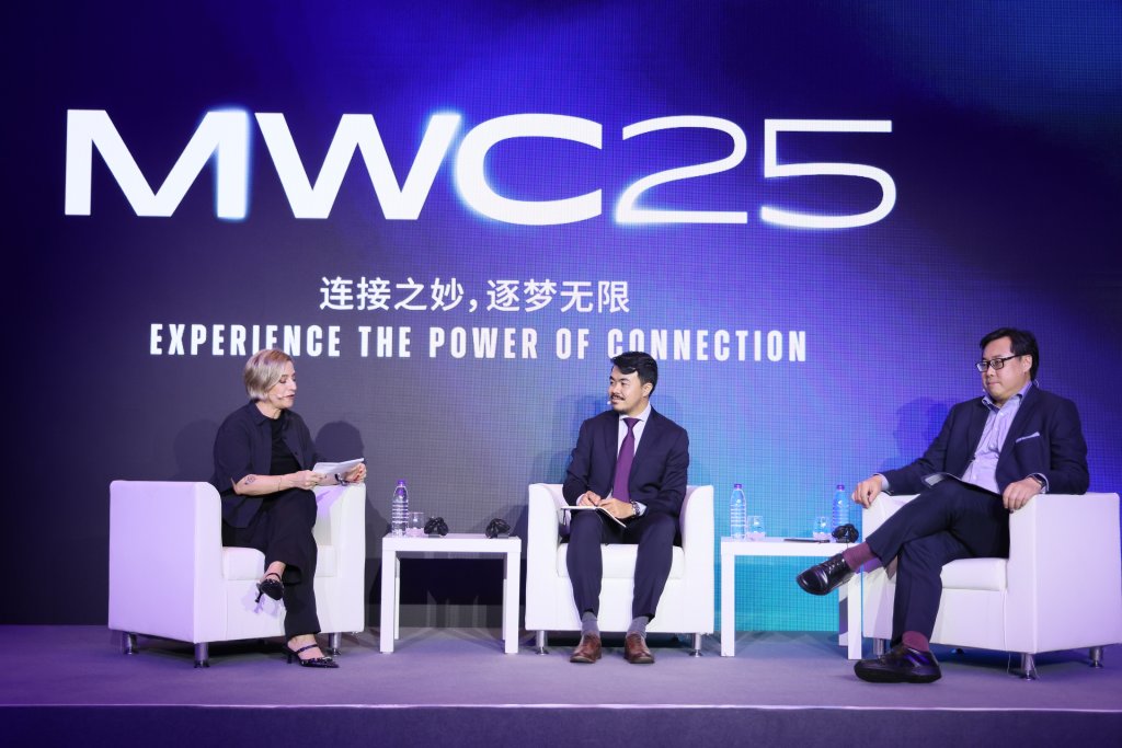 Three people sit on stage in white chairs under a large “MWC25” sign, with the tagline “Experience the Power of Connection” and Chinese characters. Two men in suits and a woman in black, holding a notepad, engage in conversation with microphones and water bottles on a table.