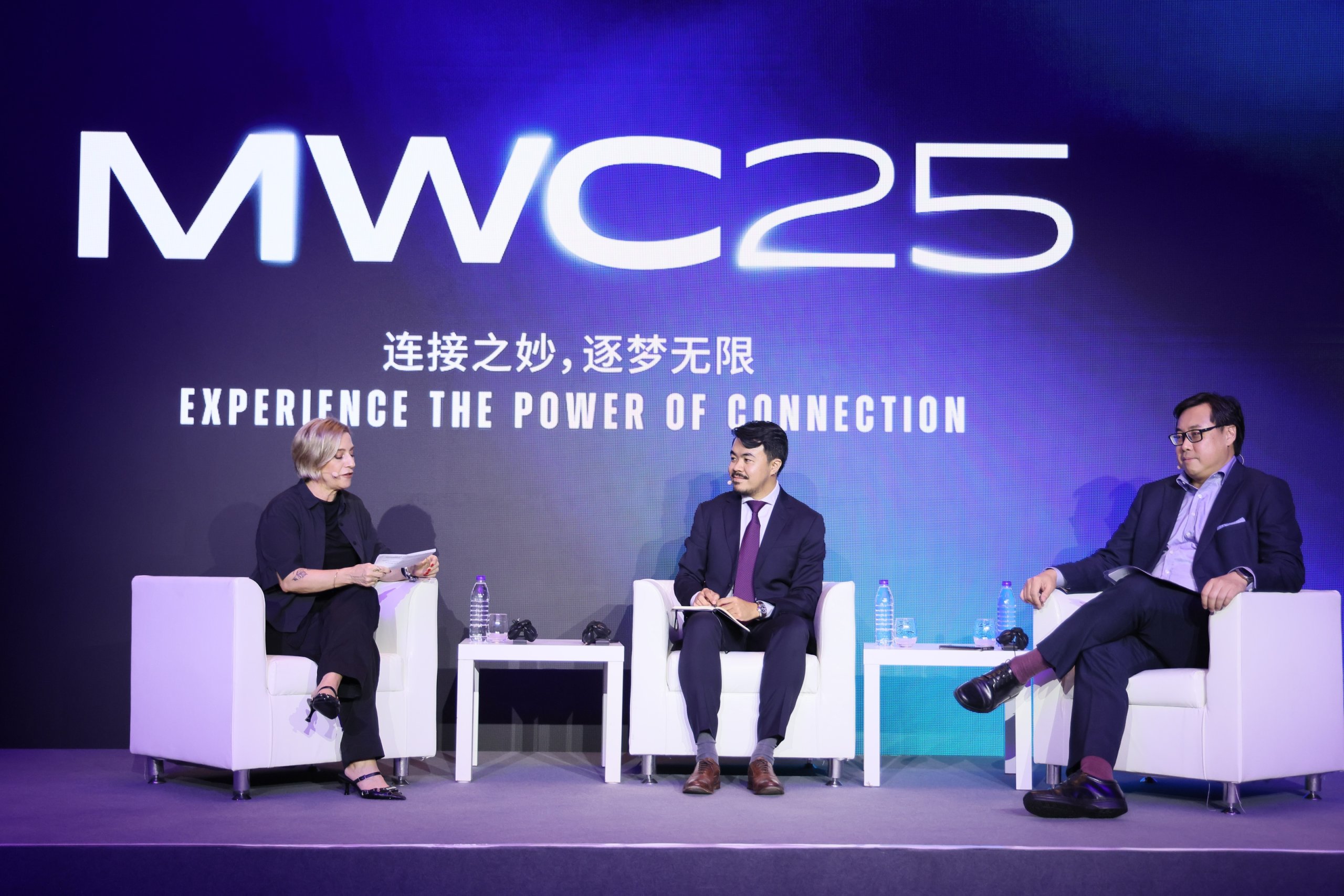 Three people sit on stage in white chairs under a large “MWC25” sign, with the tagline “Experience the Power of Connection” and Chinese characters. Two men in suits and a woman in black, holding a notepad, engage in conversation with microphones and water bottles on a table.