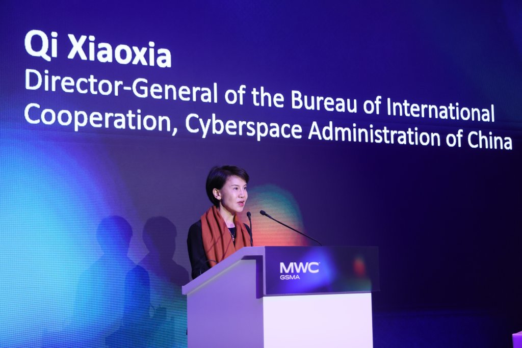 A woman stands at a podium labeled "MWC GSMA," speaking on stage. Behind her, a large screen displays her name, "Qi Xiaoxia," and her title as Director-General of the Bureau of International Cooperation, Cyberspace Administration of China.