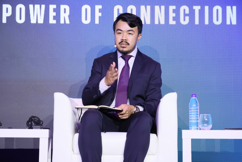 A man in a navy suit and purple tie sits on a white chair, holding an open notebook. He gestures with one hand and wears a headset mic. Behind him, large text reads “POWER OF CONNECTION.” A purple water bottle and glass are on a table beside him.