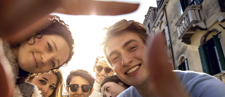 A group of six young people, smiling and posing closely together outdoors, take a selfie on a sunny day. Two hands frame the photo from the foreground, capturing youth perspectives with historic building facades in the background.