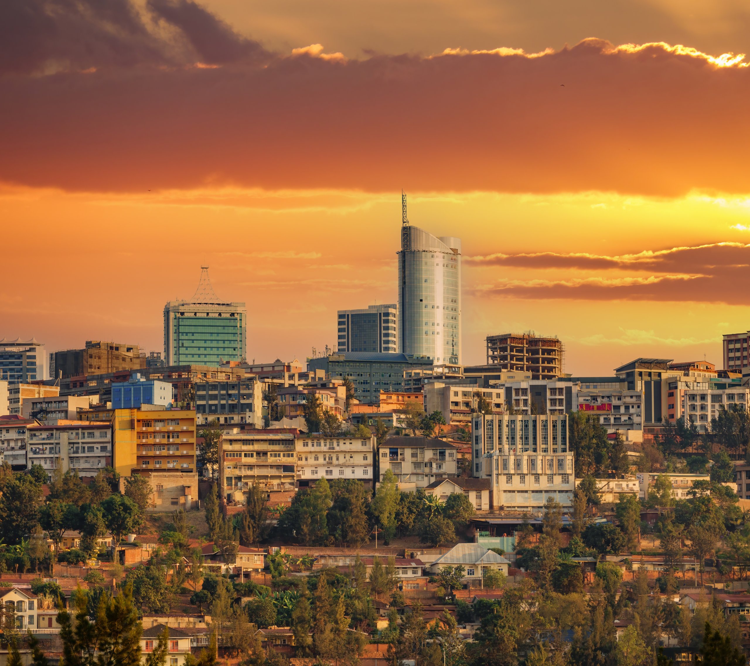 A cityscape at sunset shows modern and older buildings under a dramatic orange sky with scattered clouds. A tall cylindrical skyscraper stands out among the diverse architecture. Trees are scattered throughout the foreground.