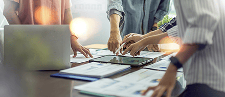 Four people stand around a table, reviewing documents and charts on smart data privacy laws, gesturing at papers and a digital tablet. A laptop is visible, suggesting a collaborative brainstorming session focused on the implementation of new policies.