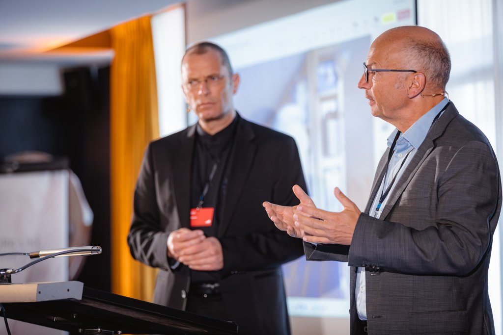 Two men in suits stand and speak at a lectern during a presentation. One man gestures with his hands whilst addressing the audience, and the other stands beside him, listening. A projected image and yellow curtain are visible in the background.